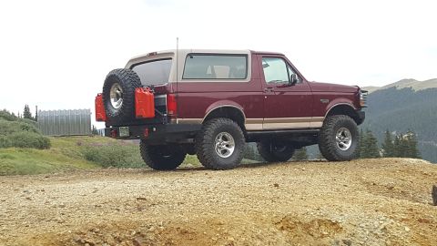 1996 Ford Bronco - The rear passenger side of a maroon 1996 Ford Bronco is shown parked on a dirt road with a mountainous, forested landscape in the backgroun…