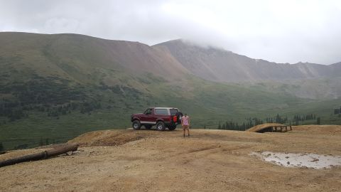 1996 Ford Bronco - A maroon 1996 Ford Bronco is parked on a dirt road with a man standing next to it. The vehicle is positioned on a ridge overlooking a vast,…