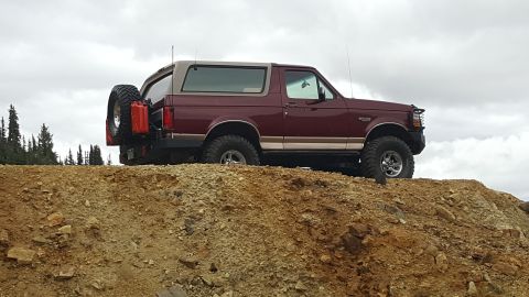 1996 Ford Bronco - A maroon and tan 1996 Ford Bronco is parked on a rocky incline with a spare tire and two red jerry cans mounted on the rear. The vehicle is…