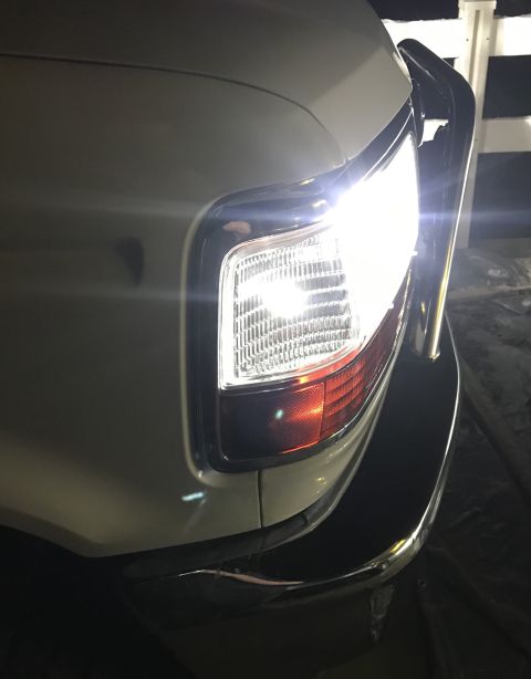 1995 Ford Bronco - Close-up of the front right headlight and turn signal assembly of a white SUV. The headlight is illuminated, casting a bright light.