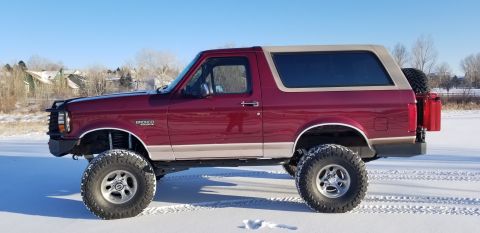 1996 Ford Bronco - A lifted, dark red 1996 Ford Bronco with a tan top is parked on a snow-covered surface under a clear blue sky. The SUV features large off-r…