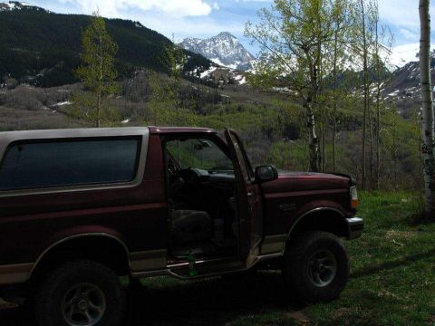 1996 Ford Bronco - A maroon 1996 Ford Bronco with the driver's side door open is parked on a grassy hill with a mountainous, wooded landscape in the backgroun…