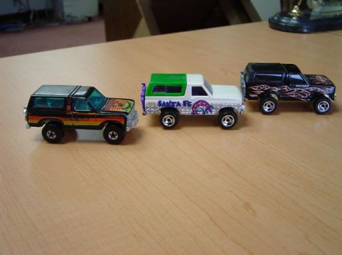 1996 Ford Bronco - Three toy Ford Bronco models are lined up on a wooden surface. The models are black with orange and yellow stripes, white with green and pu…