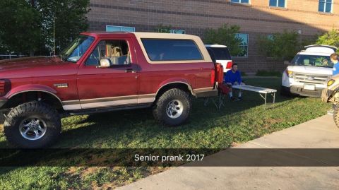 1996 Ford Bronco - A red 1996 Ford Bronco with a tan top is parked on grass next to a white SUV. A young man in a blue shirt sits on a folding table between t…