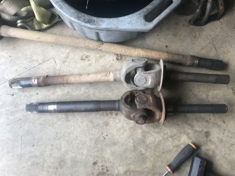 1989 Ford Bronco - Two used driveshafts with universal joints are laid out on a concrete floor. A rusty metal pan and a yellow tow strap are visible in the ba…