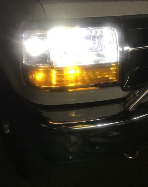 1995 Ford Bronco - Close-up of a vehicle's lit rectangular headlight with an amber turn signal glowing above a chrome bumper at night.
