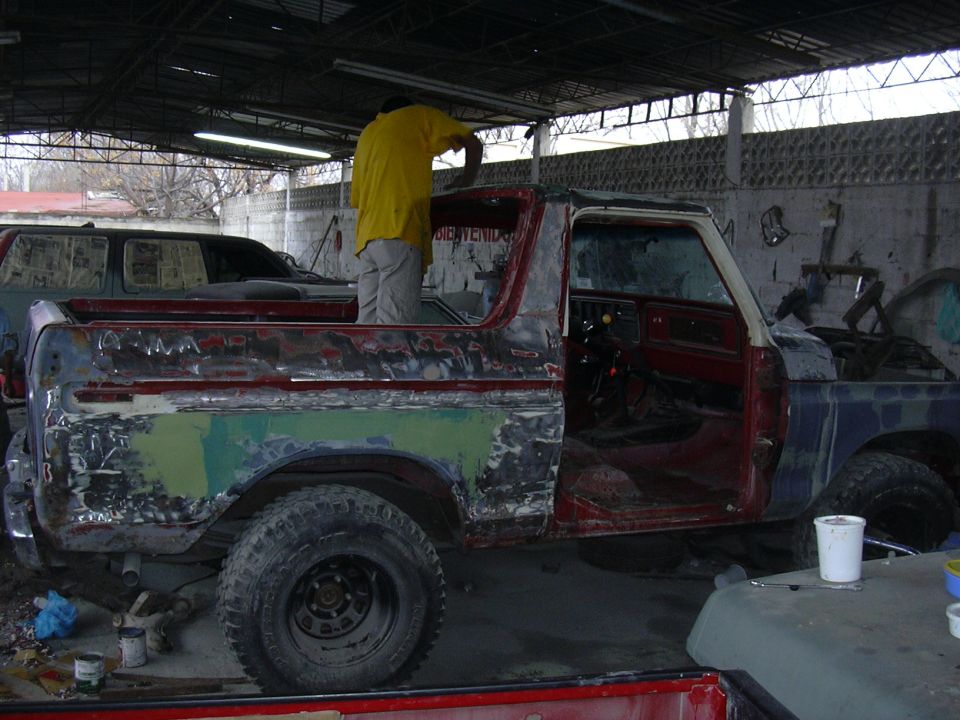 Man in yellow shirt working on a doorless Ford Bronco mid-bodywork under a covered shop