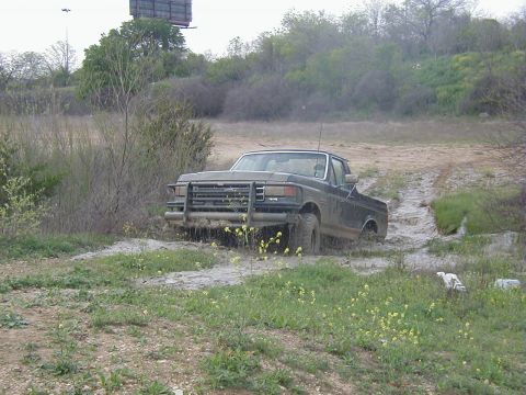 1991 Ford Bronco - Three-quarter front view of a topless dark Ford Bronco climbing out of a muddy wash, mud coating the flanks and dripping off the tires. A l…