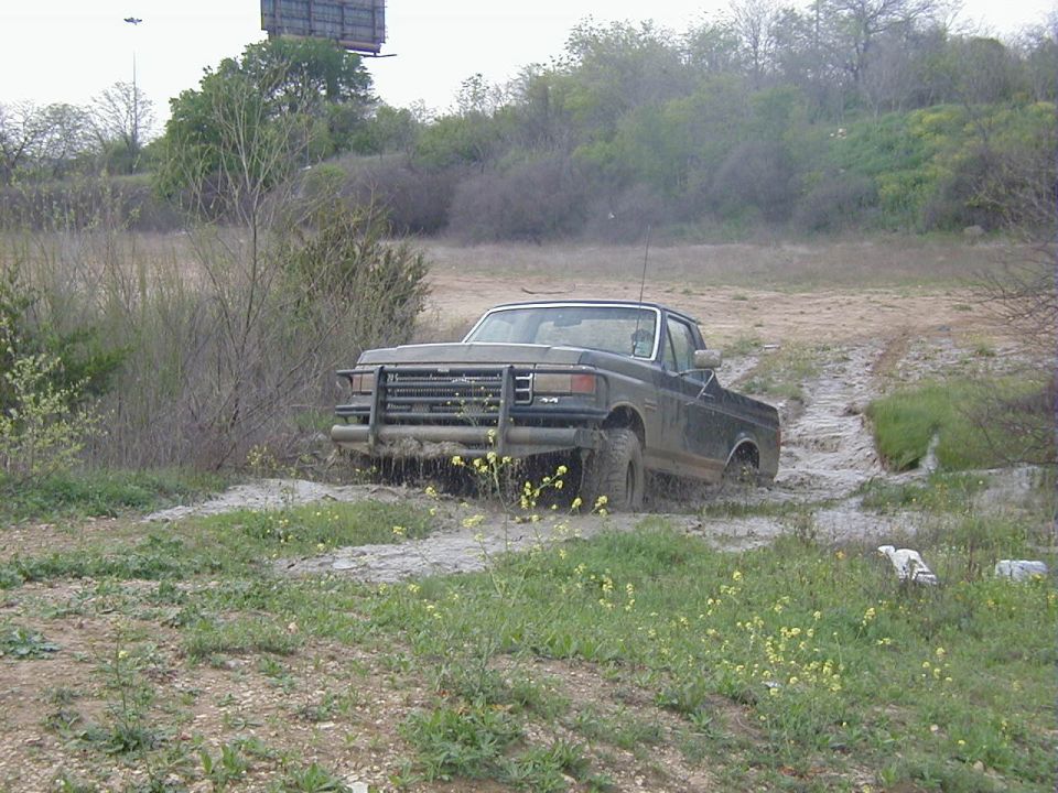 Topless mud-covered Ford Bronco with tube brush guard climbing out of a muddy wash