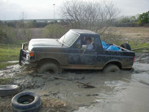 1991 Ford Bronco - Driver-side profile of a topless two-tone Ford Bronco buried to the rocker panels in a mud pit, mud splattered the length of the body and a…