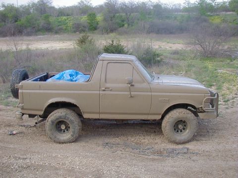 1991 Ford Bronco - Passenger-side profile of a full-size Ford Bronco with the rear top removed, coated bumper-to-roof in dried tan mud after a run. A blue tar…