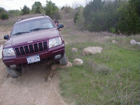 1991 Ford Bronco - Front view of a maroon Jeep Grand Cherokee flexing over a rutted dirt trail, left front wheel dropped into a washout and the body leaning.…