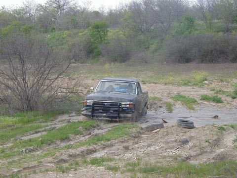 1991 Ford Bronco - Elevated three-quarter front view of a topless dark Ford Bronco sitting in a muddy pit, mud caked over the hood, fenders and tires. A tube…