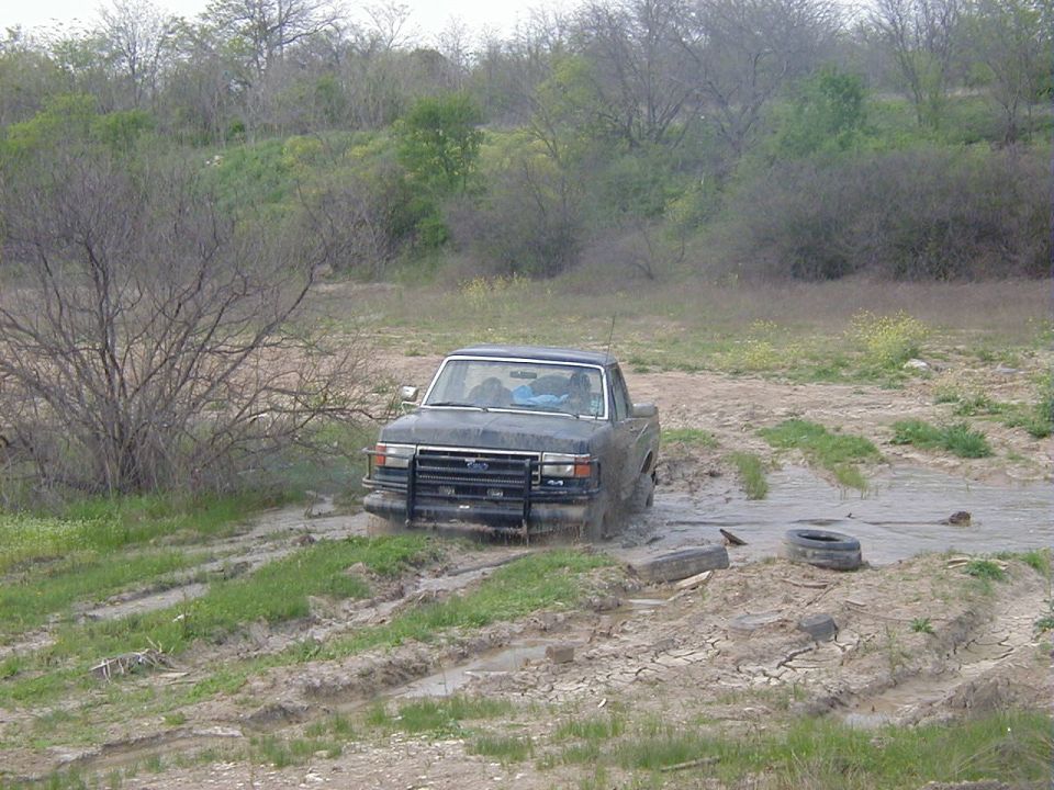 Topless mud-caked Ford Bronco with brush guard sitting in a muddy pit, two occupants