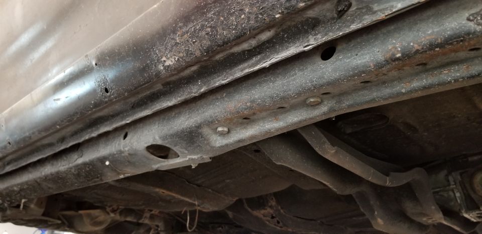Underside of a vehicle showing rusted frame rails and suspension components.