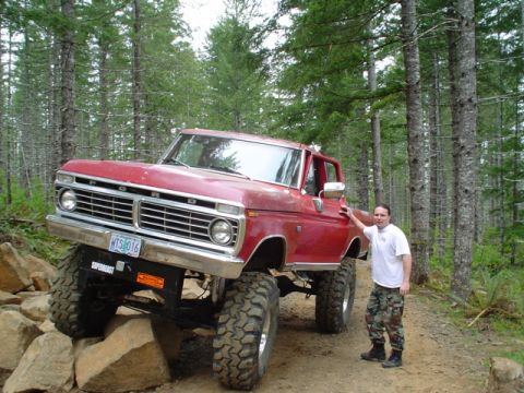 1976 Ford F-100 - A man poses with a lifted red Ford pickup truck navigating a rocky off-road path in a forest.