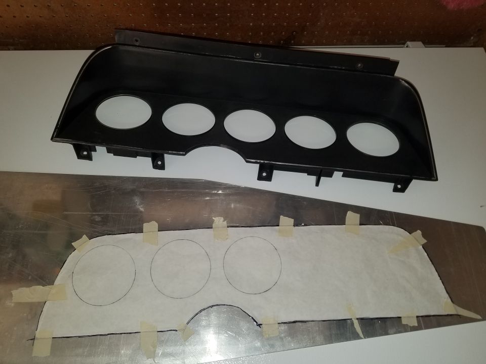 Black plastic dashboard bezel with five circular gauge openings above a metal sheet with a paper template for gauge cutouts