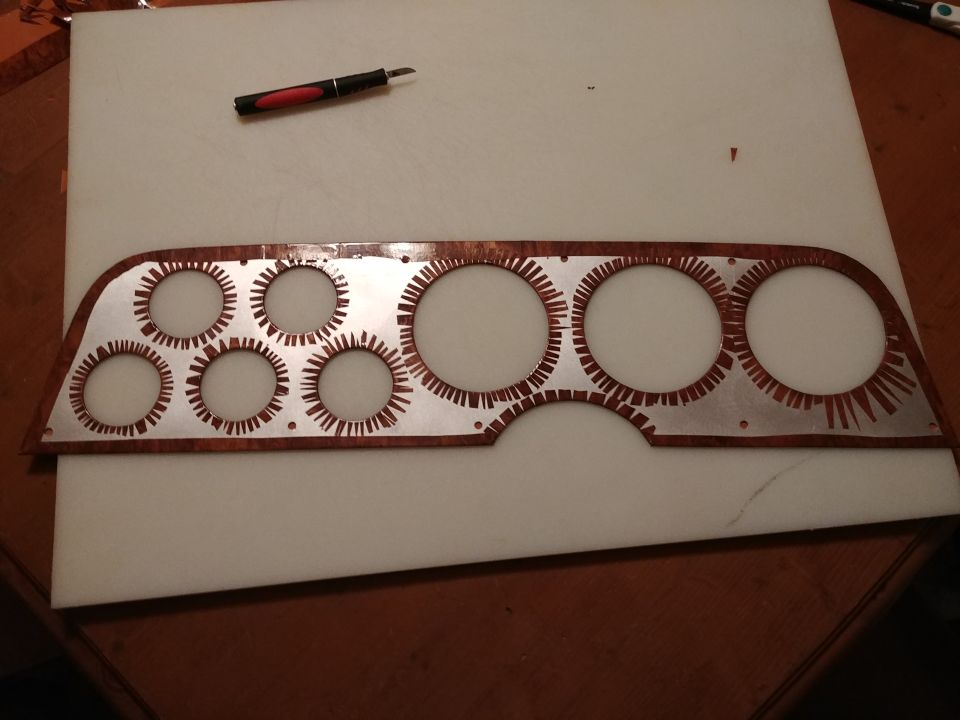 Woodgrain instrument cluster bezel with multiple circular cutouts and a craft knife on a white cutting board