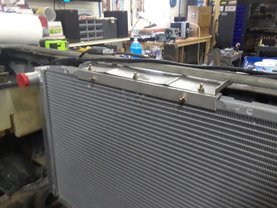 Radiator with red cap on left side, mounted in engine bay. Metal bracket above it held by bolts