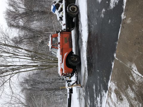 1961 Ford F-350 - An orange truck equipped with a snow plow is seen parked on a snowy road, with "GREENS" visible on the door.