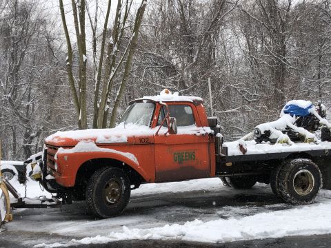 1961 Ford F-350 - A snow-covered orange flatbed truck with a snow plow is parked on a snowy road, with trees in the background.