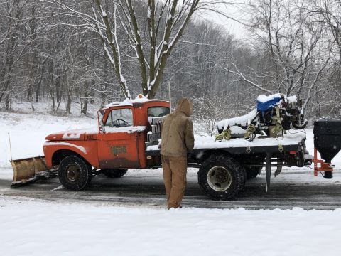 1961 Ford F-350 - A person stands next to an orange 1961 Ford F-350 truck equipped with a snow plow and a salt spreader during winter.