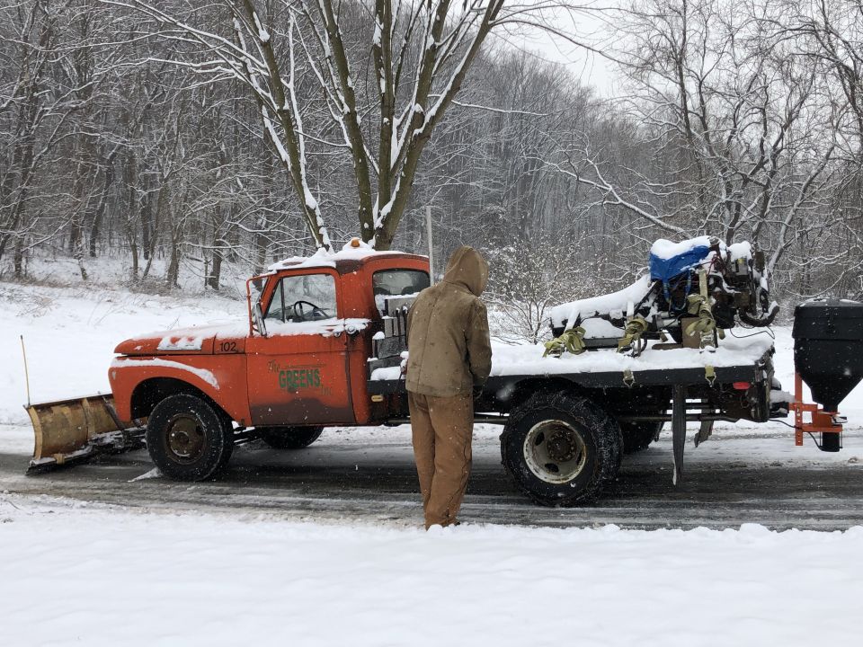 Orange 1961 Ford F-350 truck with a snow plow and a spreader on the back, parked in the snow