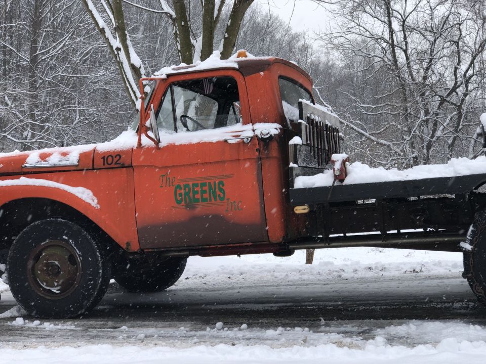 Orange flatbed truck with snow plow equipment, covered in snow. The truck has "The GREENS Inc." logo on the door and the number 102
