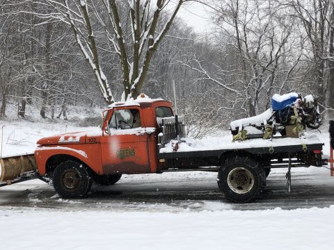1961 Ford F-350 - An orange truck equipped with a snow plow and a flatbed carrying an engine is parked on a snow-covered road.