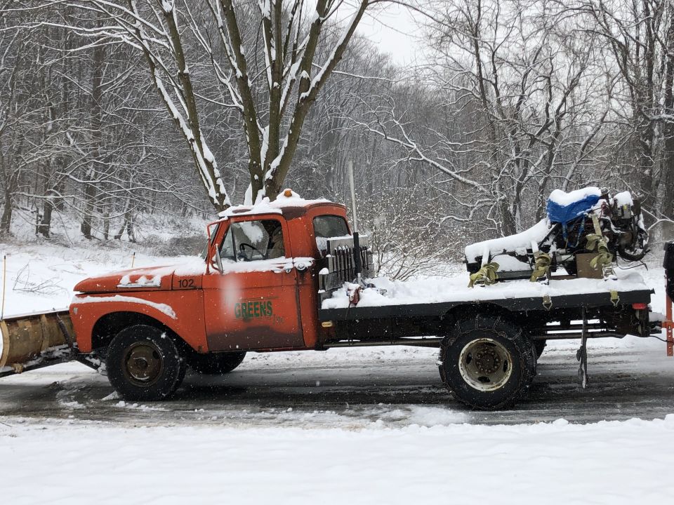 Orange truck with a snow plow attachment and a flatbed carrying an engine, in a snowy environment