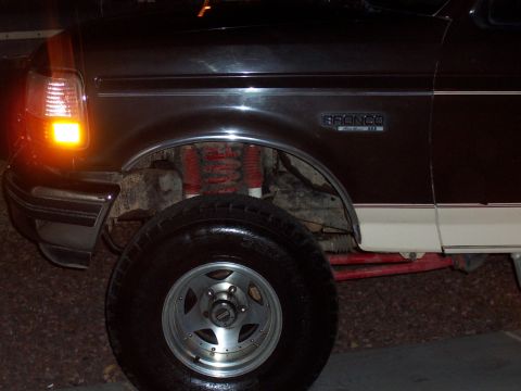 1992 Ford Bronco - Night close-up of the front wheel well of a dark Ford Bronco with Eddie Bauer badging, showing red-painted coil springs and red shock boots…