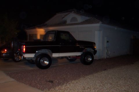 1992 Ford Bronco - Night shot of a dark full-size Ford Bronco with the hardtop removed, parked in a residential driveway beside a white stucco house with tile…