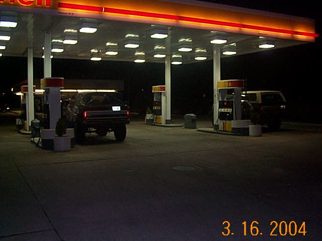 1986 Ford Bronco parked at a Shell gas station at night, with another vehicle visible in the background
