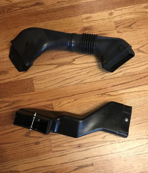 1995 Ford Bronco - Two black plastic air intake ducts are shown on a wooden floor. One is a flexible tube with a ribbed section.