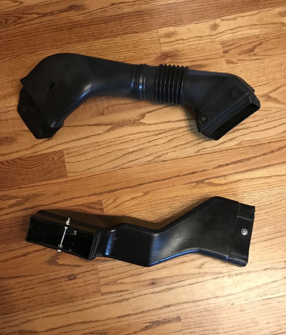 Two black plastic air intake ducts on a wooden floor. One is a flexible tube with a ribbed section