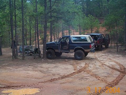 1986 Ford Bronco - Two SUVs are parked in a wooded area with tire tracks visible in the dirt.