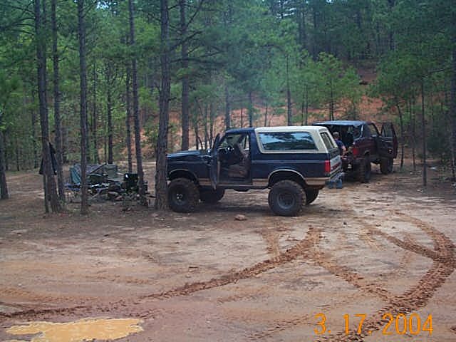 Blue Ford Bronco parked in woods with another SUV behind it. Tire tracks in the dirt