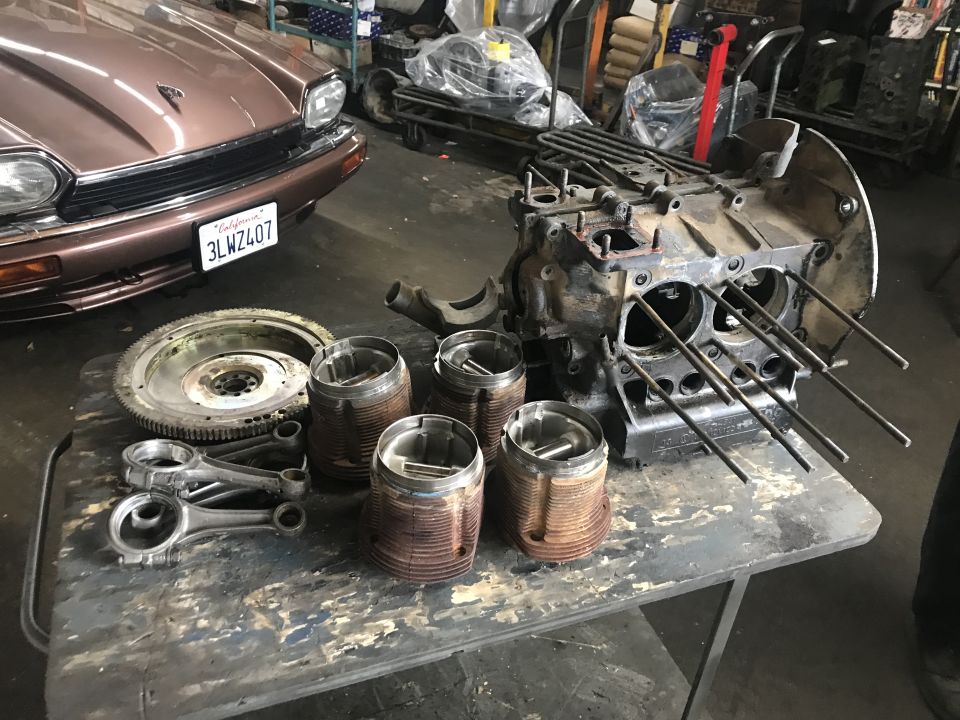 Volkswagen Beetle engine block, pistons, connecting rods, and flywheel on a table; Jaguar XJ in background
