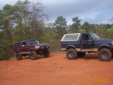1986 Ford Bronco - Two Ford Broncos are parked on a dirt path surrounded by trees under a cloudy sky.