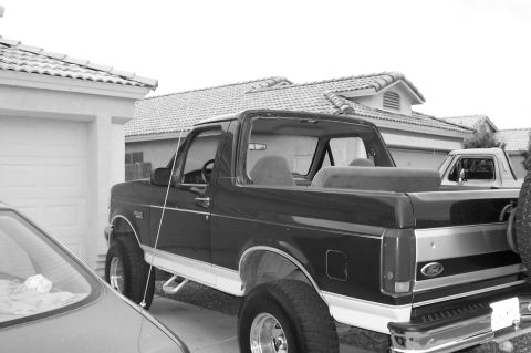 1992 Ford Bronco - Black-and-white rear three-quarter view of a lifted 1990s Ford Bronco with its fiberglass top removed, showing the exposed roll bar, bare r…