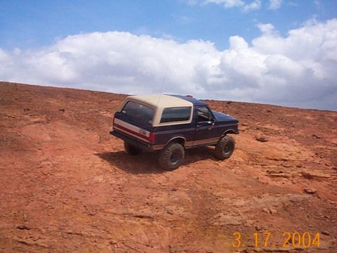 1986 Ford Bronco - Ford Bronco navigates a steep, dusty incline on a sunny day with scattered clouds.