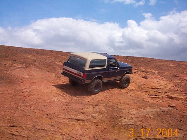 Dark blue Ford Bronco with a tan top driving on a red dirt hill under a cloudy sky