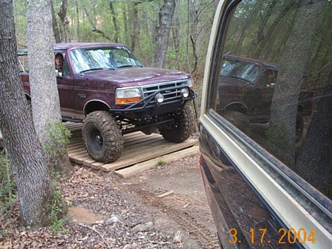 1986 Ford Bronco - A maroon Ford Bronco navigates a wooden bridge in a wooded area, with another vehicle's reflection visible in the foreground.