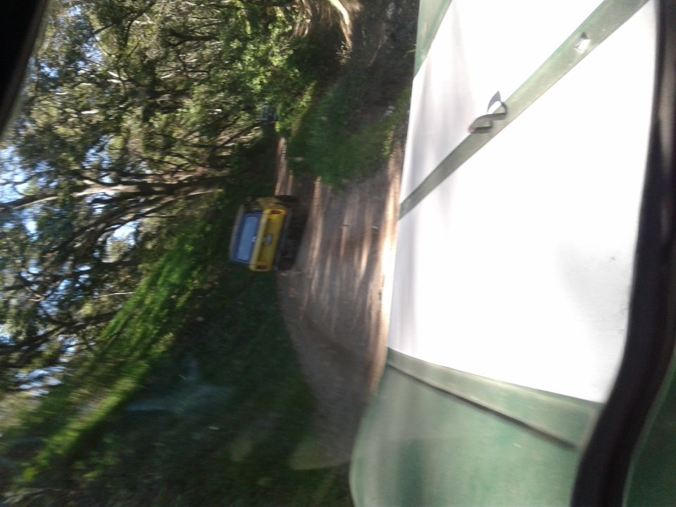 Yellow and blue vehicle on a dirt road, partially obscured by trees and a white surface