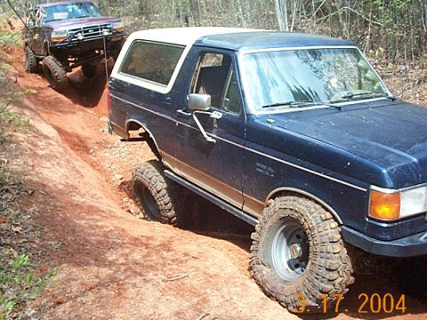 1986 Ford Bronco - Two SUVs navigate a muddy, wooded trail, with one vehicle appearing to tow the other.