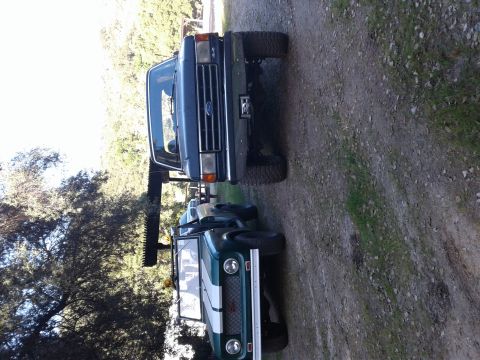 1991 Ford Bronco - Two SUVs are parked side-by-side on a dirt path, with trees in the background.