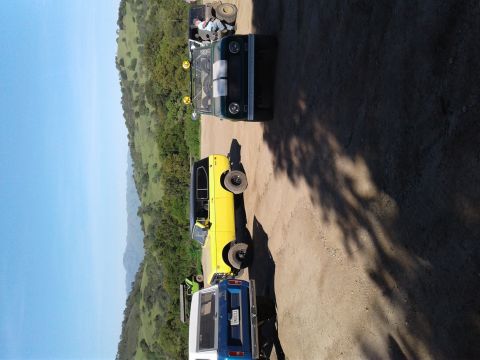 1991 Ford Bronco - Several vehicles are parked on a dirt road with a scenic backdrop of rolling green hills under a clear blue sky.
