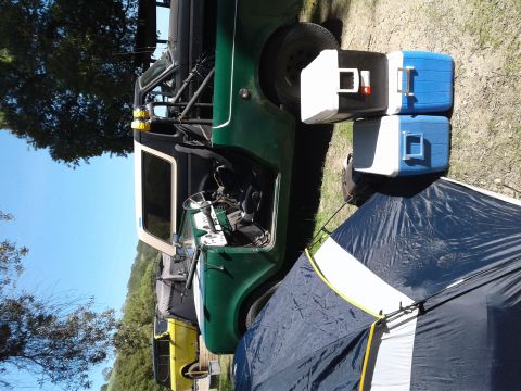 1991 Ford Bronco - A green off-road vehicle with a roll cage is parked outdoors next to coolers and a tent, with another yellow vehicle visible in the backgro…