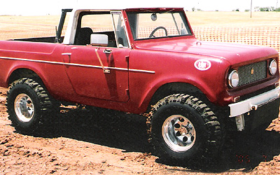1991 Ford Bronco - This red SUV, likely a 1960s International Harvester Scout, is shown from a three-quarter front angle on a sandy surface.
