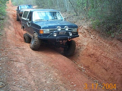 1986 Ford Bronco - Two off-road vehicles navigate a muddy, rutted trail through a wooded area.